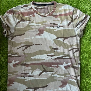 Camouflage t-shirt från Hollister - Snygg t-shirt från Hollister med klassiskt camouflagemönster i beige, brun och gröna toner. T-shirten har rund hals, korta ärmar och en broderad Hollister-logga på bröstet. Perfekt för dig som gillar streetwear och vill sticka ut med en cool look.