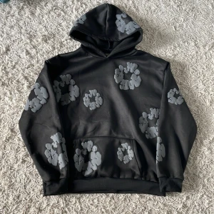 Svart hoodie med grått mönster Denim Tears - Svart hoodie från Denim Tears med unikt grått blommigt mönster över hela tröjan. Tröjan har huva, magficka och ribbade muddar vid ärmslut och nederkant. Perfekt för dig som gillar streetwear och vill sticka ut med cool design.