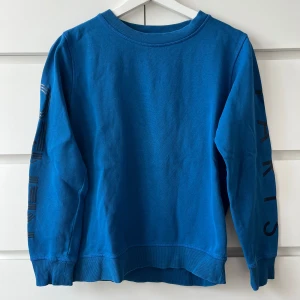 Blå sweatshirt med tryck på ärmarna - Blå sweatshirt i bomull med coolt svart tryck på båda ärmarna, där det står 'PARIS' och grafiska linjer. Passar bra för den som är 170 ungefär, sitter som en xs