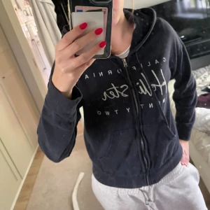 Mörkblå hoodie från Hollister - Mörkblå hoodie från Hollister med vit text framtill. Perfekt för chill dagar och har en klassisk, avslappnad passform. Materialet är mjukt och känns skönt mot huden.