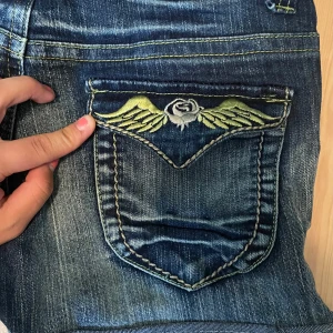 Blå jeansshorts med broderi MAS fashion - Snygga blå jeansshorts från MAS fashion med coolt broderi av vingar och ros på fickorna. Shortsen är utsydda i midjan och saknar en knapp. Jag kan posta med knappen kostnadsfritt ifall du själv vill sy fast den!  Står xl på lappen men passar som s