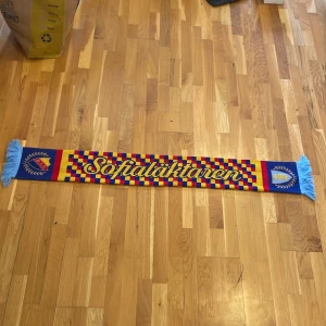 Randig Sofialäktaren halsduk DIF - Säljer en färgstark halsduk från Sofialäktaren med Djurgårdens IF-logga. Från 2023! Halsduken har ett rutigt mönster i blått, rött och gult samt ljusblå fransar i ändarna. Perfekt accessoar för att visa ditt stöd på matchen! 