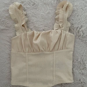 Beige linnetopp med volangband - Supersöt beige linnetopp med breda axelband dekorerade med volanger. Toppen har en lätt rynkad överdel och är tillverkad i ett strukturerat tyg som känns mjukt mot huden. Perfekt att styla med jeans eller kjol för en fräsch look.