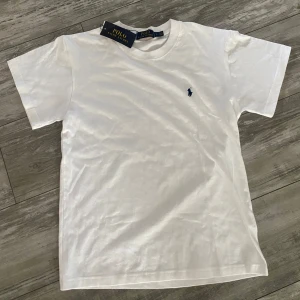 Vit t-shirt från Polo Ralph Lauren - Vit t-shirt från Polo Ralph Lauren med klassisk rund hals och korta ärmar. På bröstet finns den ikoniska blå broderade loggan. T-shirten är gjord i mjuk bomull och har en clean, stilren look som funkar till allt. Perfekt för dig som gillar enkel och snygg design helt ny med pris lapp och Ralph Lauren påse. Passar 14 åringar.