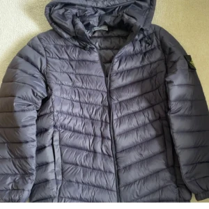 Mörkgrå dunjacka från Stone Island - Snygg mörkgrå dunjacka från Stone Island med ikonisk patch på ärmen. Jackan har huva, dragkedja framtill och quiltad design. Perfekt för kalla dagar och ger en clean streetwear-look.