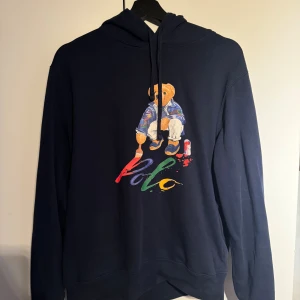 Mörkblå Polo Ralph Lauren hoodie -  mörkblå hoodie från Polo Ralph Lauren med  björnen på bröstet. Färgglad Polo-logga och  tryck med björnen som målar. huva med snörning och långa ärmar. 
