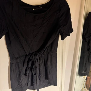 Svart knyt topp från Fire & Glory - Svart knytt top från Fire & Glory i storlek XS. Modellen har rund hals, korta ärmar och ett snyggt knytband i midjan som ger en schysst siluett. 