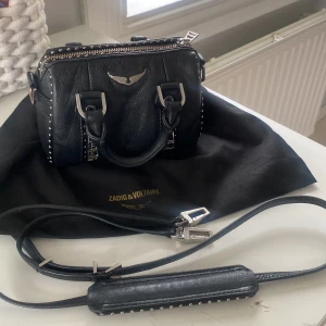 Svart handväska Zadig & Voltaire - Cool svart handväska i skinn från Zadig & Voltaire med silverfärgade metalldetaljer, nitar längs kanterna och ikonisk vinge framtill. Väskan har både handtag och avtagbar axelrem samt dragkedja upptill. Perfekt för dig som gillar edgy och stilren design.
