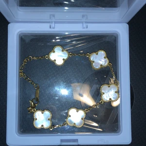 Guldarmband med vita fyrklöver - Elegant guldarmband med fem vita fyrklöverformade hängen. Varje hänge har en fin pärlemorfinish som ger en lyxig känsla. Perfekt för att lägga till en touch av elegans till din stil.