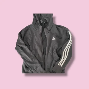 Svart huvtröja från Adidas - Säljer en svart huvtröja från Adidas med dragkedja och de klassiska tre ränderna på ärmarna. Tröjan har en huva och är långärmad, perfekt för en sportig look. Adidas-loggan syns på bröstet. Perfekt för vardagsbruk eller träning.