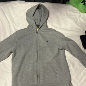 Grå hoodie från Polo Ralph Lauren - Säljer en stilren grå hoodie från Polo Ralph Lauren. Den har en dragkedja framtill och en klassisk logga broderad på bröstet. Perfekt för en avslappnad look. 🐎