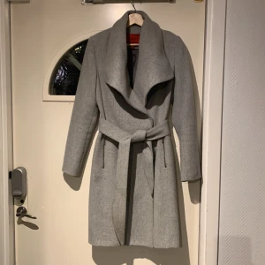 Grå kappa från Mango - Snygg grå kappa från Mango med ett elegant bälte i midjan. Kappan har en stilren design med långa ärmar och en mjuk krage. Perfekt för kyligare dagar när du vill hålla stilen. 🧥✨