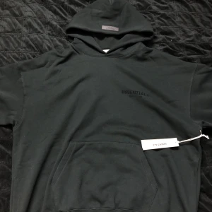 Svart hoodie från Fear of God Essentials - Snygg svart hoodie från Fear of God Essentials med logotyp på bröstet. Den har en klassisk känguruficka och en bekväm huva. Perfekt för en stilren och avslappnad look.