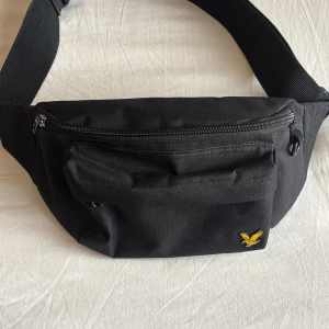 Svart Lyle&Scott  midjeväska med gul logotyp - Snygg svart midjeväska med en gul logotyp på framsidan. Väskan har flera fack med dragkedja och ett justerbart bälte för perfekt passform. Perfekt för att hålla dina saker säkra och nära till hands.