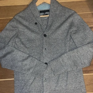 Tvärfet Grå kofta från hilfiger - Snygg grå kofta med knappar framtill och ribbade muddar. Perfekt för kyligare dagar och ger en stilren look. Passar bra till nästan allt.