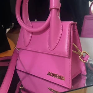 Rosa handväska från Jacquemus - Snygg rosa handväska från Jacquemus i skinn. Väskan har ett stilrent och modernt utseende med ett handtag och en axelrem. Den har en elegant guldfärgad logotyp på framsidan och detaljer i metall. Perfekt för att ge din outfit en färgklick! Äkta köpt i harrods i London 