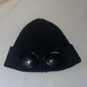 C.P. Company mössa - Snygg svart ribbad mössa från C.P. Company med unika glasögondetaljer framtill. Perfekt för att ge en cool touch till din outfit. Tillverkad i ett mjukt och bekvämt material.  Bild 2 ser man att den är lite trasig men det syns inte mycket alls.