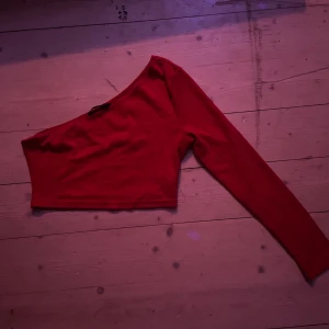 Röd oneshoulder topp - Snygg röd oneshoulder topp med lång ärm. Perfekt för en trendig look. Toppen är i en härlig röd färg och har en stilren design.