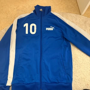 Blå träningsjacka från Puma - Säljer en blå träningsjacka från Puma med vita detaljer och nummer 10 tryckt på bröstet. Jackan har dragkedja och ribbad krage. Perfekt för sportiga aktiviteter.
