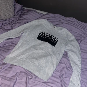 Grå sweatshirt från Hugo Boss - Säljer en grå sweatshirt från Hugo Boss med stort svart tryck på framsidan. Tröjan har långa ärmar. Passar bra till både jeans och mjukis. Perfekt för våren, priset kan sänkas kom bara privat för frågor, helt oanvänd också