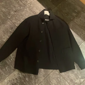 Svart kappa - Snygg svart kappa med knappar framtill. Perfekt för en stilren look. Den har en klassisk krage och raka linjer som ger en elegant siluett.