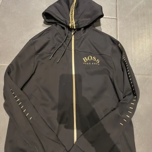 Svart zip tröja från Hugo Boss - Snygg svart hoodie från Hugo Boss med gulddetaljer. Den har en dragkedja framtill och en justerbar huva med textdetaljer. Perfekt för en stilren look.