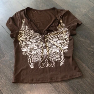 Brun t-shirt med fjärilstryck - Snygg brun t-shirt med ett stort, detaljerat fjärilstryck i vitt och guld på framsidan. T-shirten har korta ärmar och en rund halsringning med knappar. Använd fåtal gånger