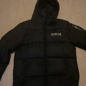 Svart pufferjacka från Hoodrich - Säljer en svart pufferjacka från Hoodrich med huva och dragkedja. Perfekt för kyliga dagar under vintern som på hösten och våren.