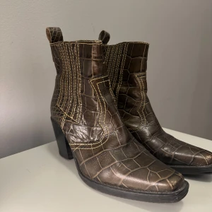 Ganni boots - Snygga bruna boots med krokodilmönster och fyrkantig tå. De har en robust klack och dekorativa sömmar som ger en unik look. Perfekta för att ge en stilfull touch till din outfit.