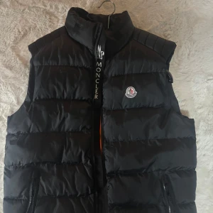 Svart dunväst från Moncler - Snygg svart dunväst från Moncler med dragkedja och logotyp på bröstet. Västen har en iögonfallande orange insida och praktiska fickor med dragkedjor. Perfekt för kyligare dagar.