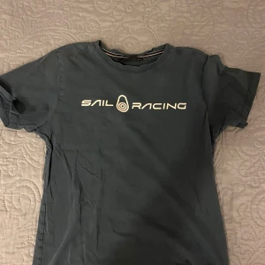 Mörkblå t-shirt från Sail Racing - Snygg mörkblå t-shirt från Sail Racing med vit logga på bröstet. Perfekt för en avslappnad stil. T-shirten har en klassisk rund hals och korta ärmar.