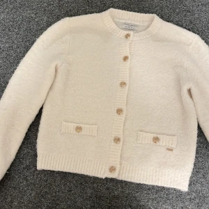 Vit kofta från Holly & Whyte storlek S - Mysig vit kofta från Holly & Whyte by Lindex med dekorativa guldknappar och två fickor framtill. Koftan har en rund halsringning och är tillverkad i ett mjukt material som ger en lyxig känsla. Perfekt för kyliga dagar.