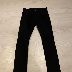 Helt nya Nudie Jeans - Snygga svarta jeansbyxor från Nudie Jeans, modell Grim Tim. Tillverkade av 99% ekologisk bomull och 1% elastan för en bekväm passform. Perfekta för en stilren look med klassisk design och knappgylf . Byxorna är helt nya utan prislapp och är i storlek 31/30
