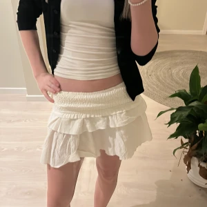 Vit volangkjol - Säljer en söt vit kortkjol med volanger. Kjolen har en elastisk midja som ger en bekväm passform. Perfekt för varma dagar och kan enkelt matchas med olika toppar.