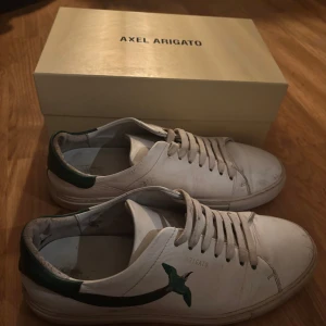 Vita sneakers från Axel Arigato - Säljer ett par vita sneakers från Axel Arigato med gröna detaljer. Skorna har snörning och är tillverkade i skinn. Perfekta för en stilren look.