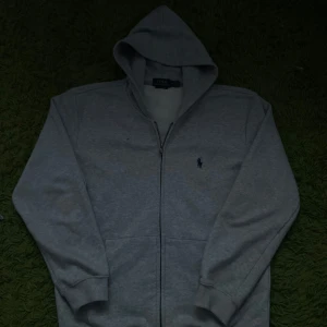 Grå hoodie från Polo Ralph Lauren - Säljer en klassisk grå hoodie från Polo Ralph Lauren med dragkedja och det ikoniska logotypbroderiet på bröstet. Perfekt för en avslappnad stil. Den har långa ärmar och en bekväm huva.