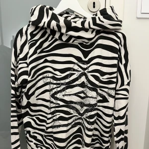 Zebramönstrad hoodie med paljettdetaljer - Snygg zebramönstrad hoodie med paljettdetaljer på framsidan. Tröjan har en bekväm passform och långa ärmar. True religion.