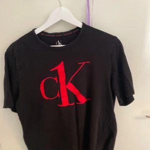 Svart t-shirt från Calvin Klein - Snygg svart t-shirt från Calvin Klein med röd logga på bröstet. Perfekt för en stilren look. T-shirten har korta ärmar och en klassisk passform.