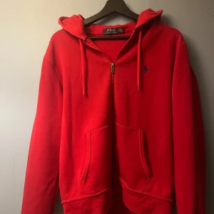 Röd Zip hoodie från Polo Ralph Lauren - Snygg röd hoodie från Polo Ralph Lauren med dragkedja och huva. Den har en klassisk design med det ikoniska logotypen broderad på bröstet. Perfekt för en avslappnad stil. ena plastbiten på snöret har lostnat men det gör ingen skillnad ändå! pris kan diskuteras vid snabb affär, säljer denna pga vill inte ha den längre för jag har tröttnat på färgen. Det är storlek M men den passar S