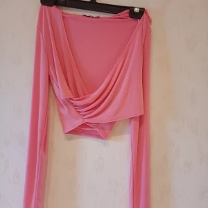 Rosa omlottopp från NLY One - Säljer en snygg rosa omlottopp från NLY One. Toppen har långa ärmar och en v-ringad design som ger en elegant look. Perfekt för att piffa upp garderoben med en färgglad touch.