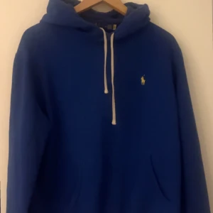 Blå hoodie från Ralph Lauren - Säljer en snygg blå hoodie från Ralph Lauren med klassisk logga på bröstet. Den har en justerbar huva med vita snören och är perfekt för en avslappnad stil. Passar bra till både jeans och joggers.