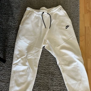 Vita mjukisbyxor från Nike - Säljer ett par vita mjukisbyxor från Nike med svart logga på sidan. Byxorna har en elastisk midja med snörning för justerbar passform. Perfekta för en avslappnad stil.