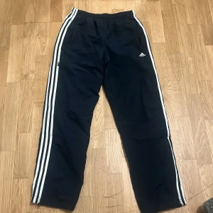 Tracksuit pant från Adidas - Snygga svarta träningsbyxor från Adidas med de klassiska tre vita ränderna längs sidorna. Byxorna har en dragkedja vid bensluten och elastisk midja för extra komfort. Perfekta för träning eller en avslappnad stil.