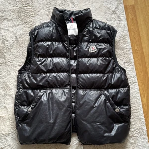 Svart dunväst från Moncler - Säljer en svart dunväst från Moncler med dragkedja och tryckknappar. Västen har en quiltad design och en broderad logotyp på bröstet. Perfekt för kyligare dagar. Inuti finns en unik etikett med serietidningsmotiv. Liten flaw på baksida. Självklart äkta. Size 2 (medium)