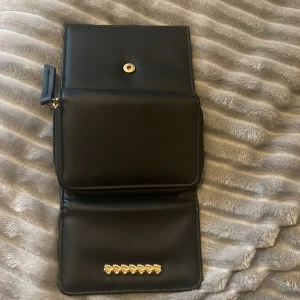 Svart plånbok med gulddetaljer - Snygg svart plånbok med gulddetaljer och praktisk dragkedja. Den har flera fack för kort och en transparent sektion för ID. Perfekt för att hålla ordning på dina tillhörigheter med stil.