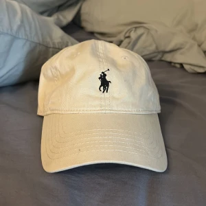 Beige keps från Polo Ralph Lauren - Snygg beige keps från Polo Ralph Lauren med den ikoniska broderade loggan framtill. Kepsen har en justerbar rem baktill för perfekt passform. Perfekt för en avslappnad stil.