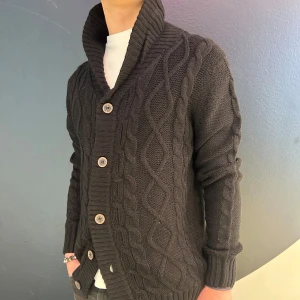 Svart stickad kofta med knappar - Snygg svart stickad kofta med ett flätmönster och stora knappar framtill. Den har en ribbad krage och långa ärmar, perfekt för kyligare dagar. Passar bra över en t-shirt för en avslappnad stil.