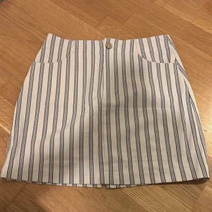 Randig kjol från Pull&Bear - Snygg kortkjol från Pull&Bear i vitt med blå ränder. Kjolen har en knapp framtill och praktiska fickor. Perfekt för en stilren look.