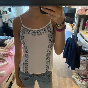 Vit linnetopp med blått mönster - Säljer en söt vit linnetopp med smala axelband och blått broderat mönster längs sidorna. Perfekt för varma sommardagar. Toppen har en enkel och stilren design som passar till det mesta. Reglerbara band💕