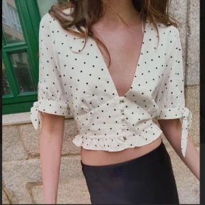 Vit prickig blus - Säljer en söt vit blus med svarta prickar och v-ringning. Blusen har korta ärmar med knytband och en volangdetalj vid midjan. Perfekt för en somrig look!